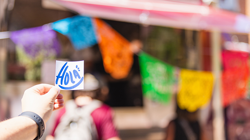 Colorful banner with hand holding sticker that says "Hola"