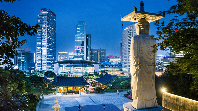 A temple statue looms in the foreground against a backdrop of modern skyscrapers