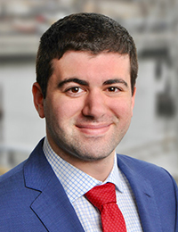 Headshot of a dark-haired man in a blue suit with white shirt and red tie.