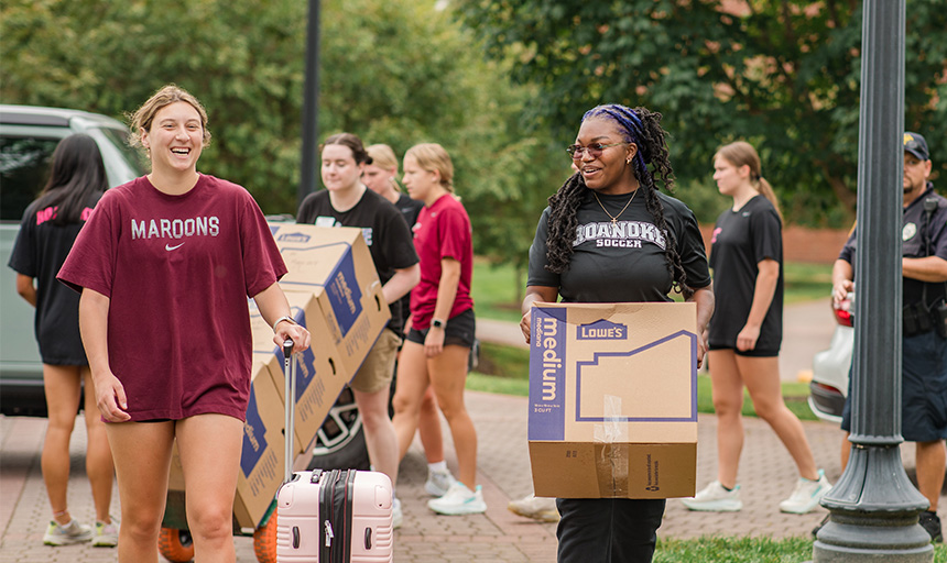 Two students smile while carrying boxes and luggage