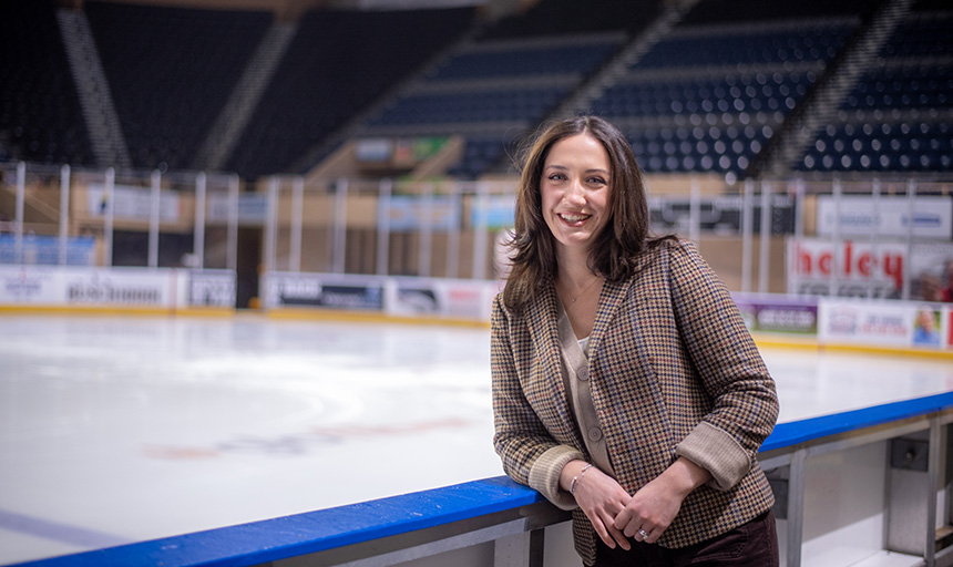 Woman leans against a railing with an ice hockey rink behind her
