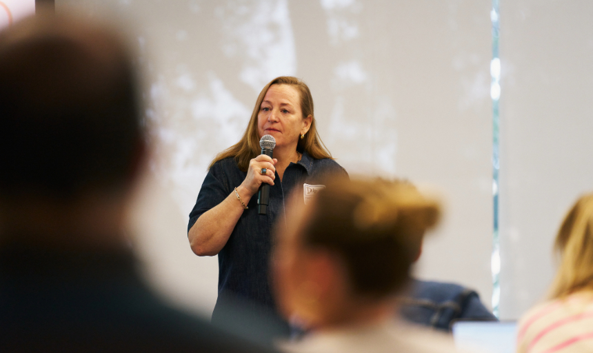 Deb (Murray) Lemon '90 holds a microphone and speaks to a group of people. 