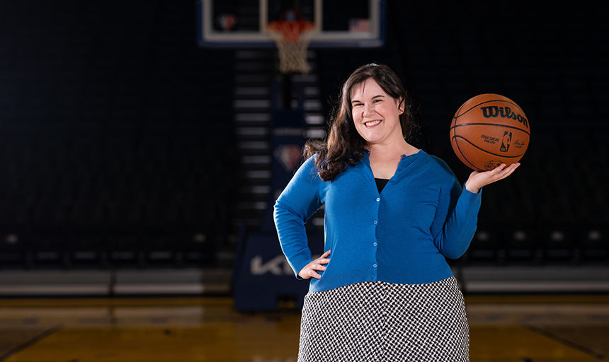 Smiling woman in blue sweater holds up a basketball on a basketball court.