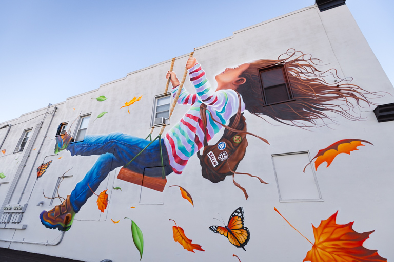 Mural on side of a white building depicting a girl on a swing, her hair blowing behind her and leaves and butterflies floating around her.