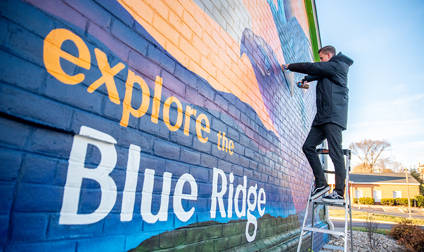 Man on a ladder spray paints a blue and purple mural with the words "explore the Blue Ridge" on it. 