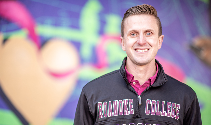Young man in a Roanoke College shirt stands in front of a purple, green and pink mural.
