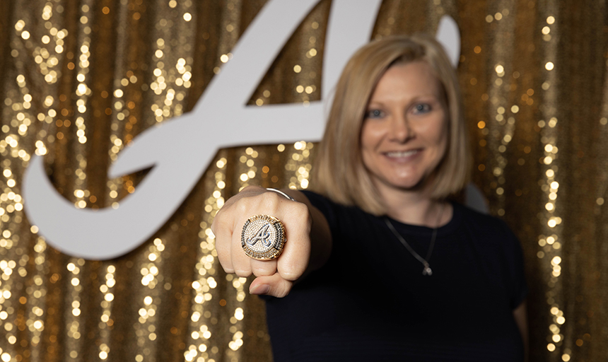 A smiling woman holds out her fist to show a large World Series ring on her finger, and she is out of focus in the background.
