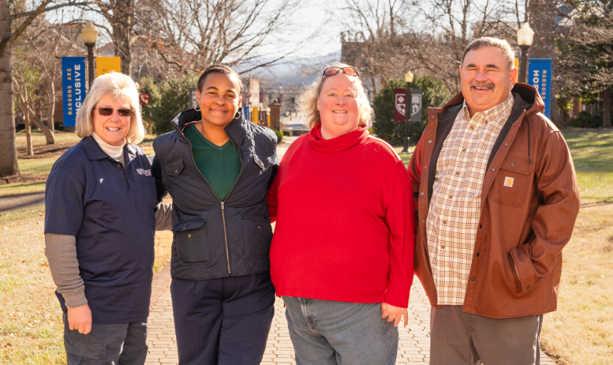 Four people, three women and a man, stand side-by-side smiling with the college campus behind them.