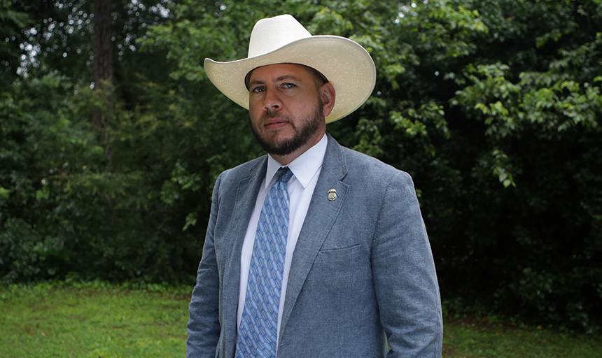 Man standing among greenery with a blue suite jacket, blue tie, khaki pants and cream colored cowboy hat