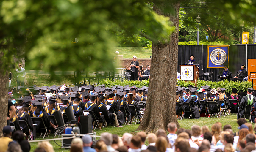 A sea of graduates clad in black gowns in rows of chairs on a green lawn with a large tree in the foreground.
