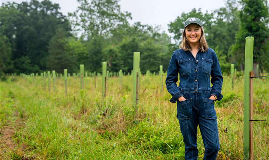 Photo of young woman in denim overalls standing in front of a green field that has rows of tree seedlings in the background. 
