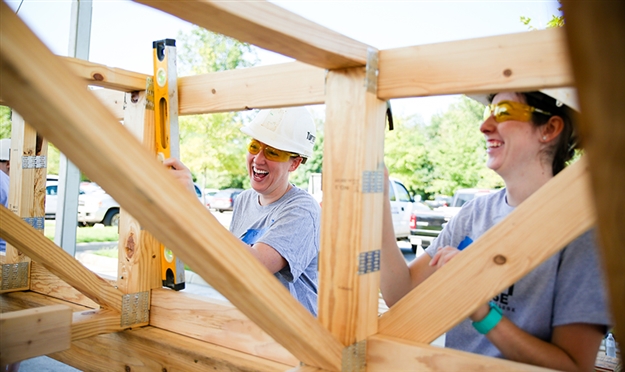 Students wearing hard hats smile while working on a house build