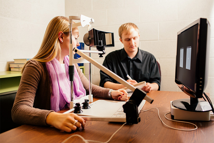 A student takes notes while another student prepares to lean into an eye device for an experiment
